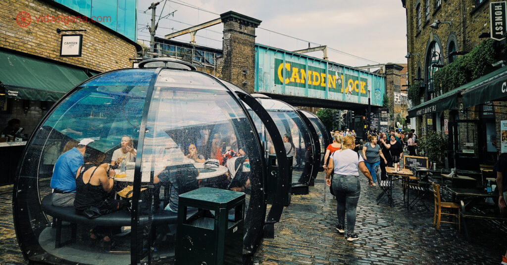 O bairro Camden, em Londres, é conhecido por seus mercados e também pelo cenário musical. A foto mostra parte da rua, em frente ao Camden Lock, onde pessoas podem comer em uma estrutura de acrílico que permite estar ao ar livre e protegido da chuva, ao mesmo tempo.