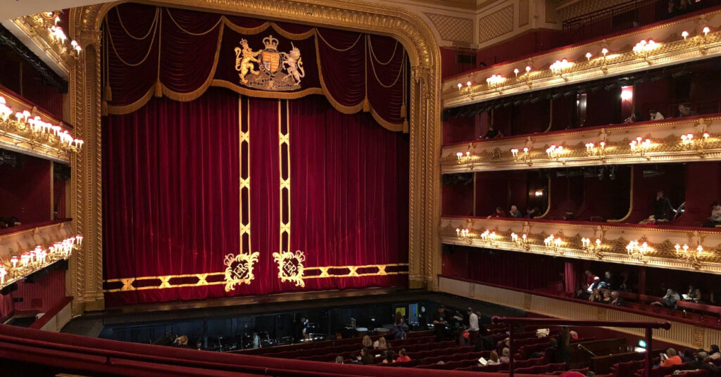 O interior da casa de óperas é luxuoso. A foto mostra o palco, fechado com cortinas carmim e detalhes em dourado. 