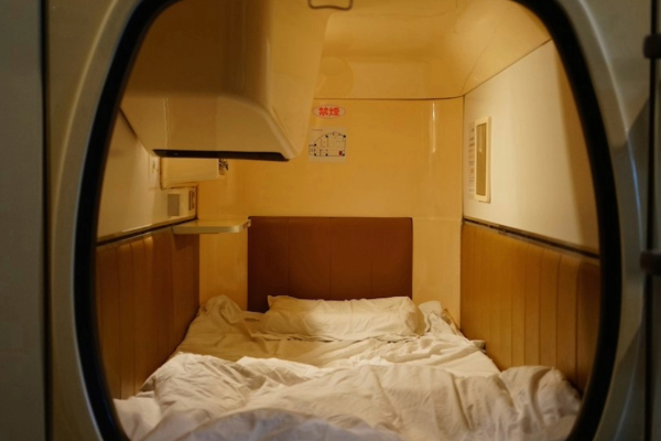 A foto mostra um exemplo de hotéis cápsula. É um compartimento bem pequeno, com apenas itens básicos para dormir.