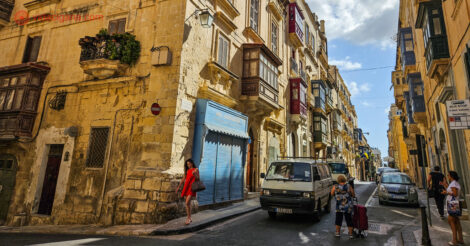 Aluguel de carro em Malta: um cruzamento em Malta com carros na rua e uma mulher vestida de vermelho andando na rua