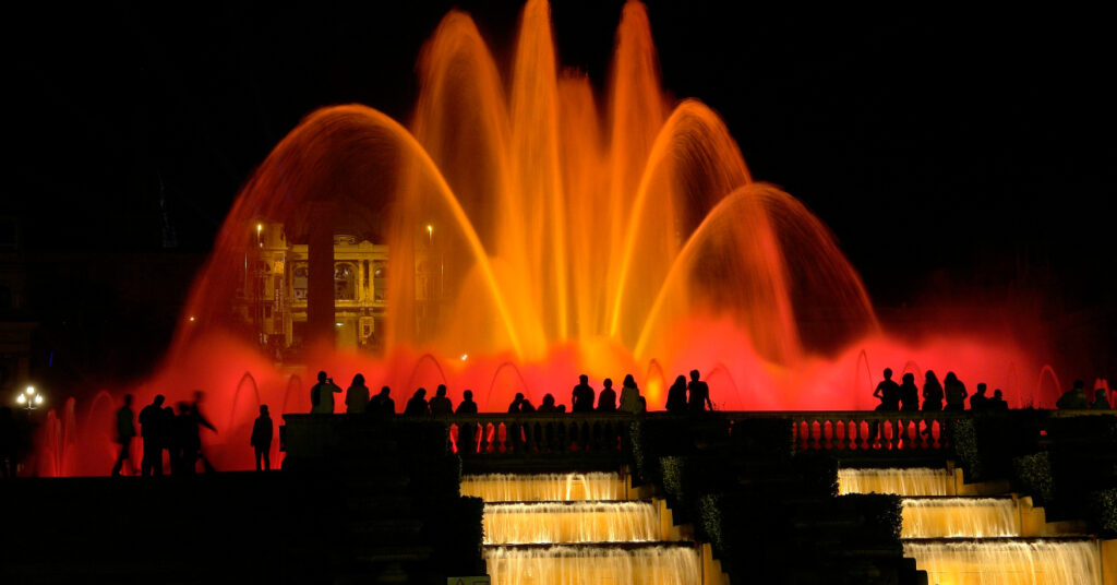 Na imagem, pessoas se agrupam em volta da Fonte Mágica de Montjuïc para assistir ao show noturno de luzes e água. 