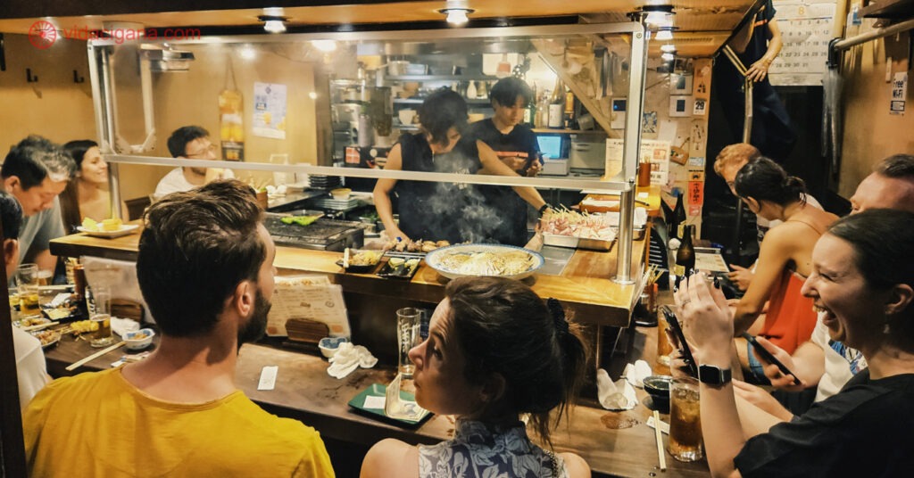 Várias pessoas dentro de um pub em Omoide Yokocho, sentadas num balcão ao redor de 2 homens cozinhando