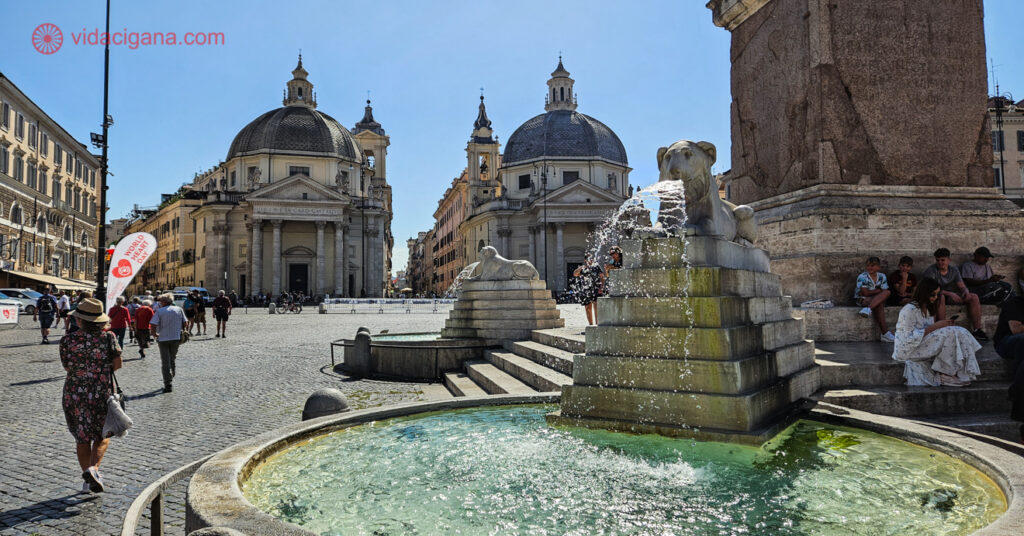 A Piazza del Popolo com as igrejas irmãs ao fundo e uma fonte com um leão soltando água pela boca em primeiro plano