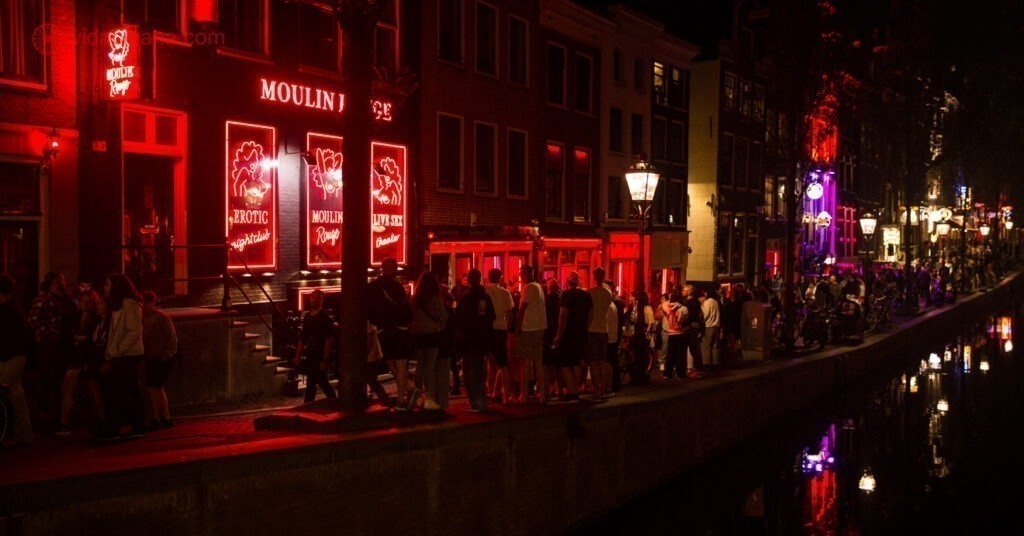 A foto mostra uma das ruas do Red Light District lotada de turistas. A região em Amsterdam é conhecida, e nomeada, pela iluminação vermelha mostrada na imagem.
