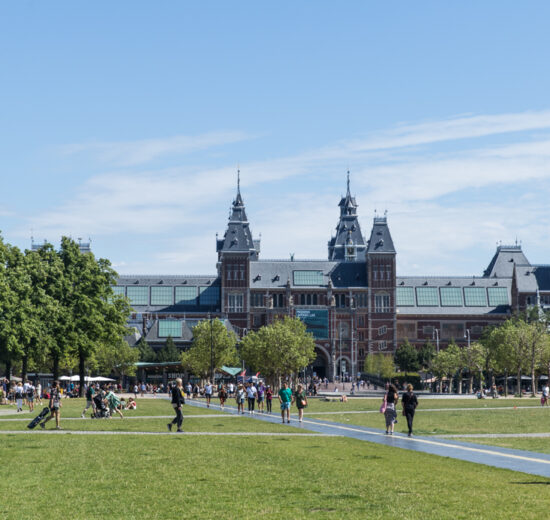 A Museumplein Amsterdam, com o Rijksmuseum ao fundo e o Museu Van Gogh do lado esquerdo. Um grande gramado é visto