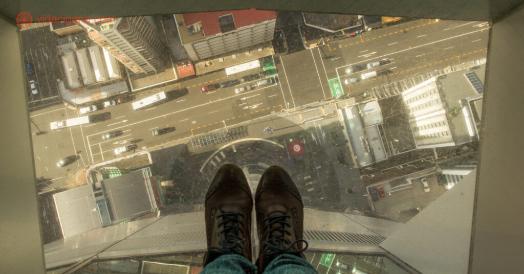 A cidade de Auckland vista do alto da Sky Tower, onde só os pés da fotógrafa aparecem, em cima de um chão de vidro de onde é possível ver as ruas e os carros lá embaixo.