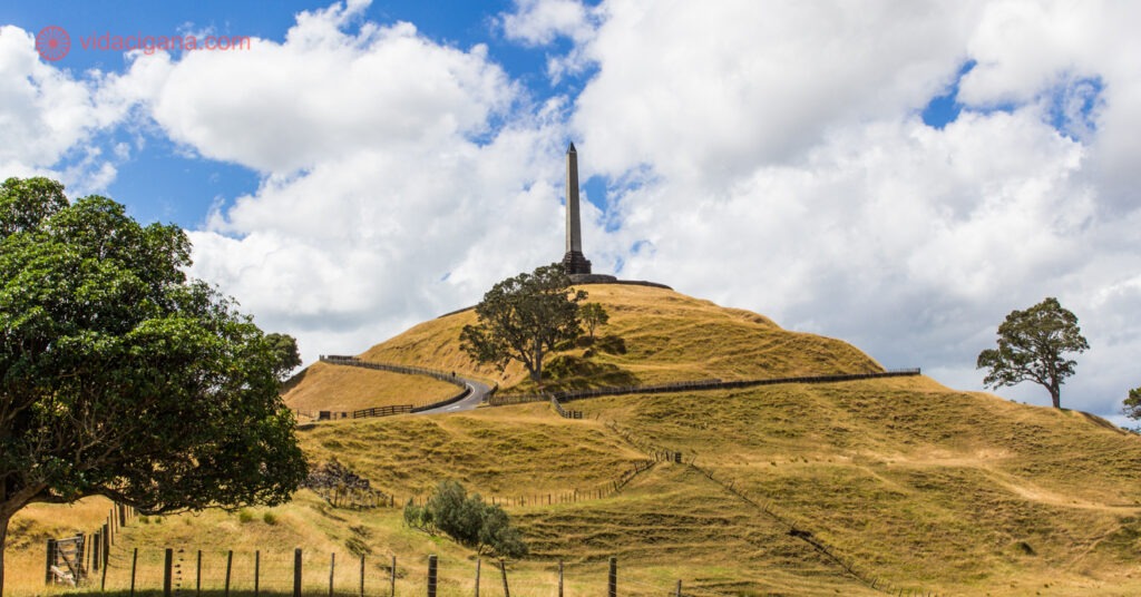 One Tree Hill, um antigo vulcão que fica em Auckland, na Nova Zelândia. A colina possui um obelisco em seu topo e um caminho que vem até seu topo. A colina está coberta de grama seca numa coloração amarelada. O céu está azul com nuvens brancas. Árvores também são vistas no caminho. Na borda da foto é vista uma cerca de madeira.