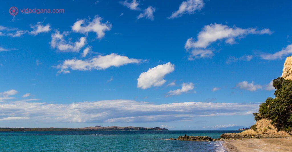 Uma pequena praia na Nova Zelândia, com uma pequena faixa de areia, com o mar claro, algumas rochas, um pedaço de terra ao fundo. O céu é azul com poucas nuvens brancas.
