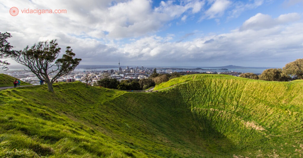 O Mount Eden, o vulcão mais famoso de Auckland, a maior cidade da Nova Zelândia. Na foto vemos o vulcão extinto com sua cratera coberta de grama verde. Uma sombra se projeta para dentro da cratera. Uma árvore se encontra na borda da cratera. O céu está parcialmente nublado, com o canto direito azul e a esquerda com bastante nuvens. Ao fundo vemos a cidade de Auckland, com a Sky Tower, a maior torre do país.