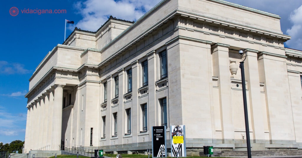 O Auckland Museum, um dos maiores museus da Nova Zelândia. Ele possui arquitetura clássica, com a fachada cheia de colunas e quinas pontudas. O céu está azul com algumas nuvens brancas.