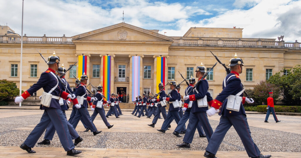 A guarda real em frente a sede da República da Colômbia. Na fachada, estão penduradas as bandeiras do país, a bandeira LGBTQIAPN+ e a bandeira do orgulho Trans. 