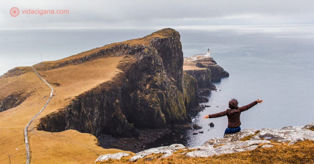 Eu na beira de uma rocha com o farol da ilha de Skye ao fundo, na ponta de uma península