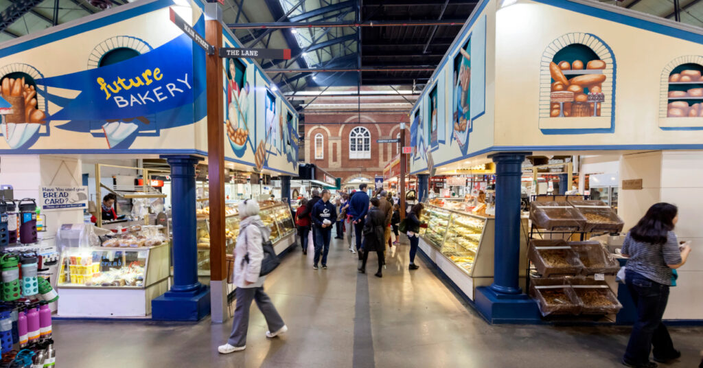 Na imagem, pessoas passeiam entre os estandes de uma padaria, no St Lawrence Market. 