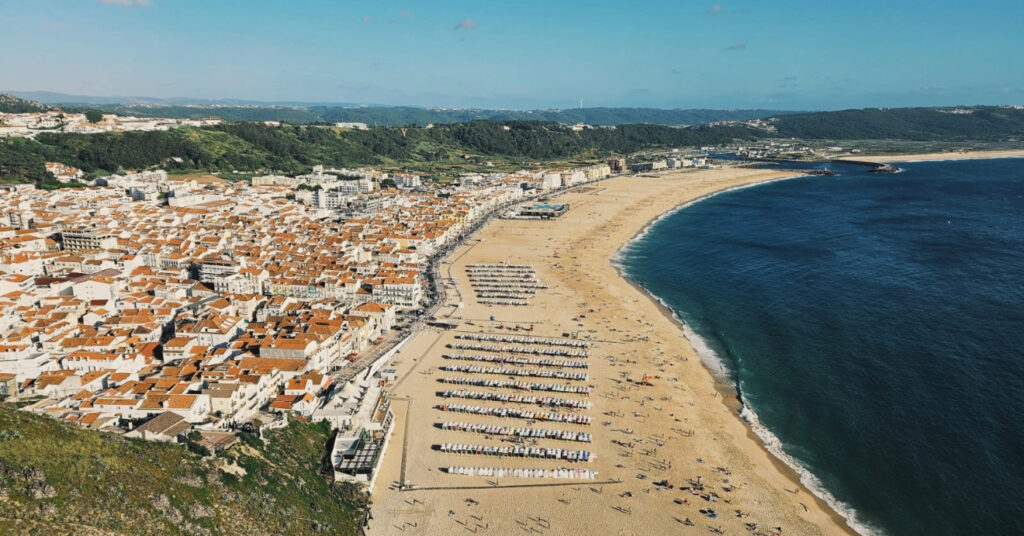 Vista panorâmica da Praia de Nazaré, seu mar calmo, faixa de areia e parte da vila. 