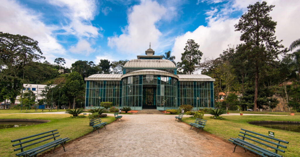 Entrada e estrutura do Palácio de Cristal, em Petrópolis. Ao redor, há um jardim, lagos e bancos. 