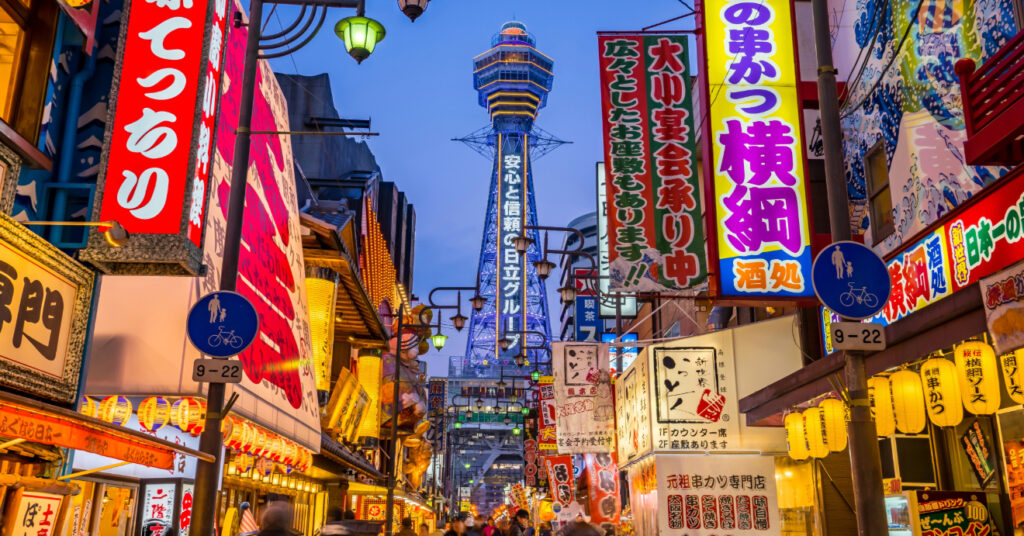 Os letreiros iluminados em uma das ruas de Shinsekai. É possível ver a Torre Tsutenkaku ao fundo. 