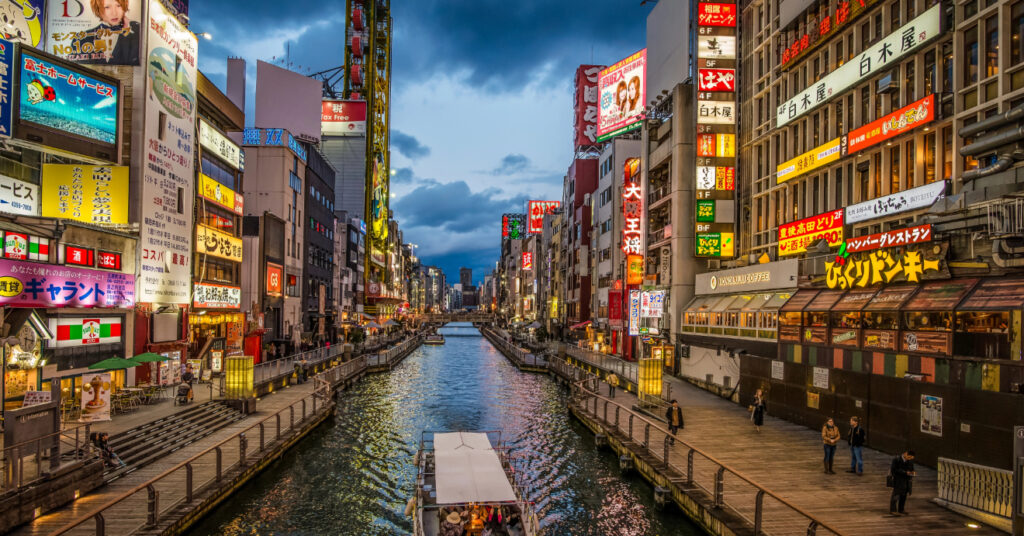 Pequena embarcação atravessa o Rio Dotonbori durante o pôr do sol. Às margens, comércio, restaurantes e bares com seus letreiros iluminados. 