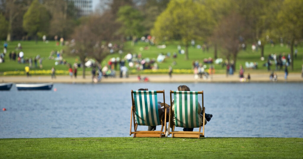 Em frente ao lago, duas cadeiras de descanso e, ao fundo, pessoas descansam e aproveitam o Hyde Park, um dos parques públicos em Londres. 