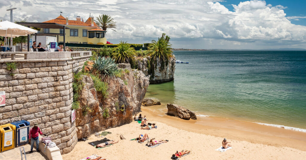 Alguns banhistas tomam sol na areia da Praia da Rainha, ao lado do paredão de pedra que delimita sua extensão. 